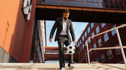 Ein Mann im Geschäftsanzug und in Turnschuhen steht auf einer Freitreppe und jongliert einen Fußball mit seinem Fuß. Er steht vor einem modernen Gebäude mit großen Fenstern und einer überdachten Fußgängerbrücke.