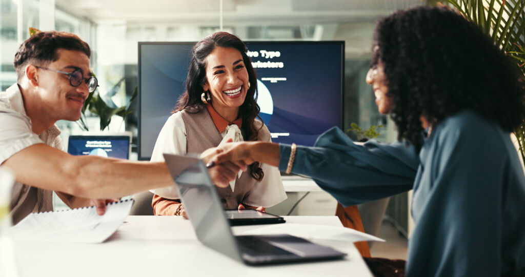 Drei Personen sitzen an einem Tisch in einem Büro. Zwei Frauen schütteln sich die Hände und lächeln, während ein Mann, der Papiere in der Hand hält, lächelnd zusieht. Laptops und Dokumente sind zu sehen, und im Hintergrund läuft eine Präsentation auf einem Bildschirm.