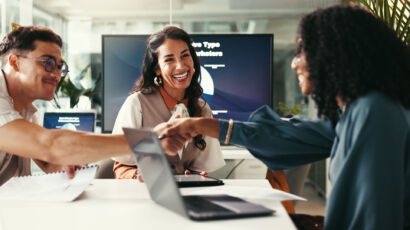 Drei Personen sitzen an einem Tisch in einem Büro. Zwei Frauen schütteln sich die Hände und lächeln, während ein Mann, der Papiere in der Hand hält, lächelnd zusieht. Laptops und Dokumente sind zu sehen, und im Hintergrund läuft eine Präsentation auf einem Bildschirm.