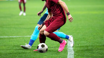 Zwei Fußballspieler in Aktion auf einem Feld, einer in einem kastanienbraunen Trikot mit rosa Schuhen und der andere in einem blauen Trikot. Der Spieler in Kastanienbraun dribbelt den Ball in der Nähe einer weißen Seitenlinie.