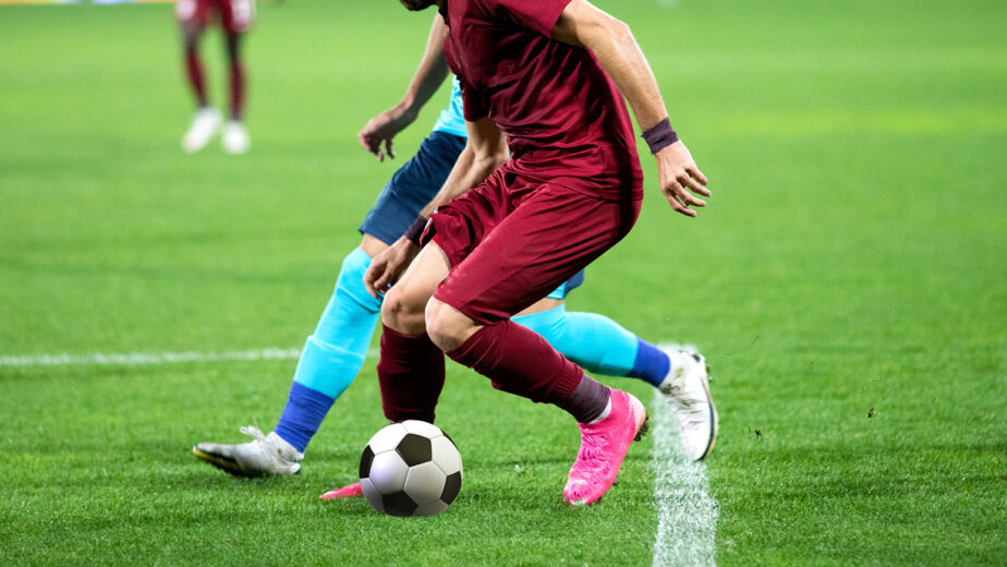 Zwei Fußballspieler in Aktion auf einem Feld, einer in einem kastanienbraunen Trikot mit rosa Schuhen und der andere in einem blauen Trikot. Der Spieler in Kastanienbraun dribbelt den Ball in der Nähe einer weißen Seitenlinie.