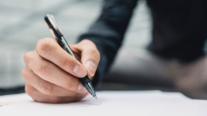 Eine Person hält einen schwarzen Stift in der Hand und schreibt auf ein weißes Blatt Papier, vielleicht macht sie sich als Spielerberater Notizen. Die Person trägt ein langärmeliges, dunkles Hemd und konzentriert sich auf ihre Hand und ihren Stift; der Hintergrund bleibt unscharf.
