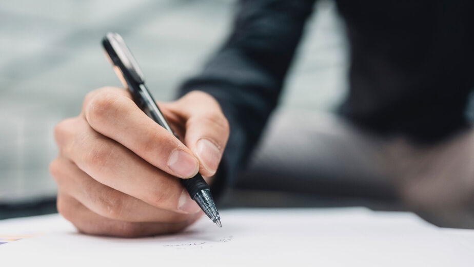 Eine Person hält einen schwarzen Stift in der Hand und schreibt auf ein weißes Blatt Papier, vielleicht macht sie sich als Spielerberater Notizen. Die Person trägt ein langärmeliges, dunkles Hemd und konzentriert sich auf ihre Hand und ihren Stift; der Hintergrund bleibt unscharf.